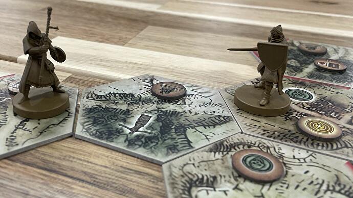 Auf einem Holztisch werden zwei kleine Figuren auf sechseckige Spielweltplättchen gestellt. Es gibt auch ein paar Tokens auf ihnen. Es sieht so aus, als hätte jemand eine tolle Brettspielzeit.
