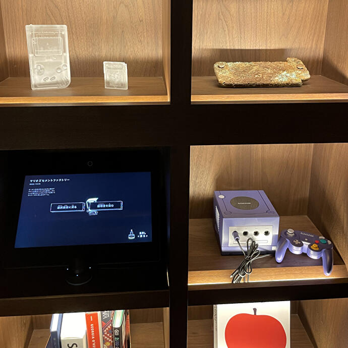 Ein Bücherregal mit etwas, das wie ein goldverkrusteter Nintendo Switch aussieht, und ein anderes mit einem GameCube darin.